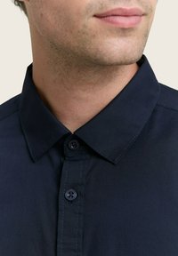 Primo piano della parte inferiore del viso e del collo di un uomo che indossa una camicia azzurra scura con colletto a punta.