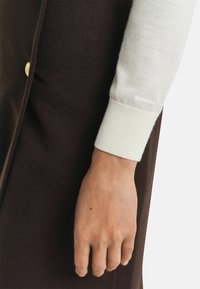 Hand und Handgelenk, das einen cremefarbenen Pulloverärmel mit einem gerippten Bund trägt, neben einem dunkelbraunen Kleidungsstück mit einem sichtbaren goldenen Knopf.