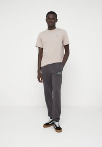 Ανοιχτό μπεζ κοντομάνικο μπλουζάκι με μικρό λογότυπο, συνδυασμένο με σκούρους γκρι φόρμες που διαθέτουν τιρκουάζ κεντήματα και μαύρα αθλητικά παπούτσια.