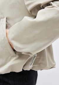 Bež jopica z dolgimi rokavi z vidnim stranskim žepom in tkanino Marc O’Polo, nošena preko temnih hlač.