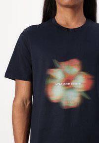 Námornícka modrá bavlnená tričko s farebnou, rozmazanou kvetinovou grafikou a textom "LYLE AND SCOTT" umiestneným v strede pod dizajnom.