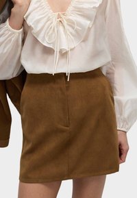 Bruine suède minirok met zijzakken en een gladde textuur, gecombineerd met een witte blouse met een rucheshalslijn en trekkoorddetail.