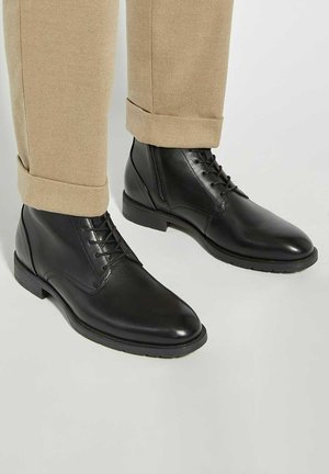 Black leather lace-up ankle boots with side zipper worn with beige cuffed trousers on white background.