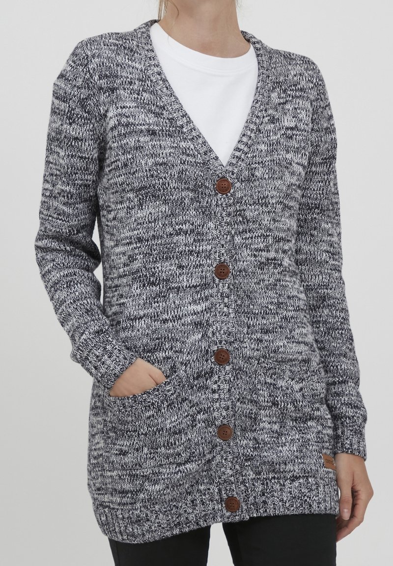 Cardigan en tricot chiné gris et blanc avec des boutons marron, poches avant, porté sur une chemise blanche et associé à un pantalon noir.
