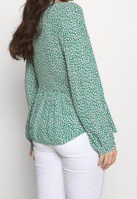Blusa floreale verde con vita a peplum, maniche lunghe e un motivo in tessuto testurizzato, abbinata a pantaloni bianchi aderenti.