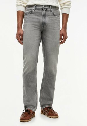 Mann trägt hellgraue gerade Jeans, braune Leder-Schnürschuhe und einen weißen strukturierten Pullover, steht mit entspannten Händen.