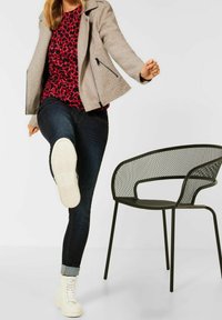 Světle šedá texturovaná bunda, červený top s leopardím potiskem, tmavé džíny, bílé vysoké tenisky. Černá kovová židle v pozadí.