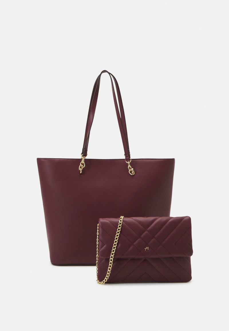 Burgundin värinen nahkakassi, jossa on kaksi pitkää kantohihnaa, yhdistettynä pienempään tikkaukseen varustettuun clutch-laukkuun, jossa on kultaketju ja hillitty logo.