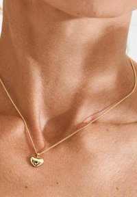 Gouden hartvormige hanger aan een fijne ketting. De ketting is dun, met een glad oppervlak en een gepolijste afwerking, rustend op een gebruinde hals.