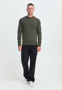 Man standing wearing olive green knit sweater, black pants, and white sneakers with black stripes on plain light background.