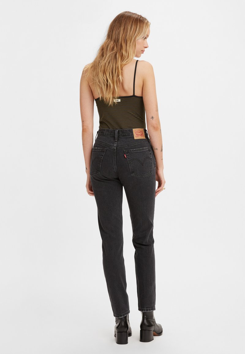 Schwarze High-Waist-Jeans mit schmaler Passform, mit Gesäßtaschen und einem Leder-Logo-Patch. Das Model trägt ein gestreiftes Tanktop und Knöchelstiefel.