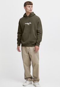 Felpa verde oliva con stampa grafica bianca, tasca a marsupio frontale, polsini e orlo ribattuti. Abbinata a pantaloni beige chiaro e sneakers bianche.