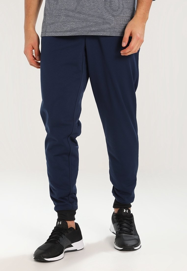 Man klädd i marinblå tapered byxor, svarta sportskor och en grå skjorta, stående mot en vit bakgrund.