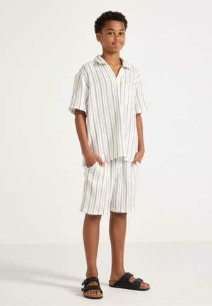 Junge steht, trägt ein helles, vertikal gestreiftes Kurzarmhemd und passende Shorts mit schwarzen Sandalen, die Hände in den Taschen.