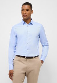 Lichtblauwe langemouwen shirt met een button-down kraag, textuurstof en witte knopen, gecombineerd met beige broek en een donkere riem.
