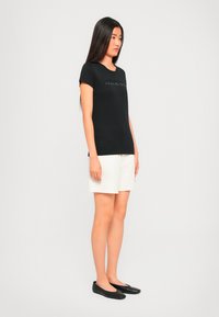 Ung kvinne som står, iført en svart t-skjorte med tekst, hvite shorts og svarte leiligheter, foran en enkel lys bakgrunn.