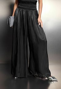 Pantalons noirs à pinces avec une taille froncée, associés à une pochette argentée texturée et des chaussures argentées brillantes à bout pointu.