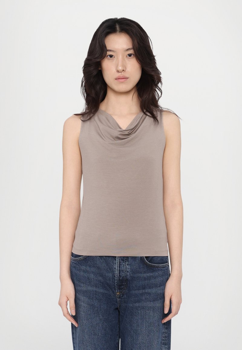 Beige, ermeløs topp med drapert hals, laget av myk fabric. Designet er tettsittende med minimale sømmer, kombinert med mørkeblå jeans.