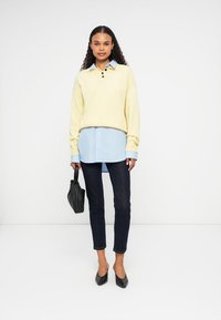 Suéter amarillo de punto con cuello, superpuesto a una camisa de rayas azul, acompañado de unos jeans oscuros, un bolso negro y tacones negros puntiagudos.