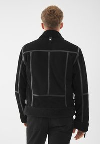 Giacca in suede nera con colletto in pelle di pecora, caratterizzata da strisce verticali, cuciture a contrasto e una finitura morbida e testurizzata.