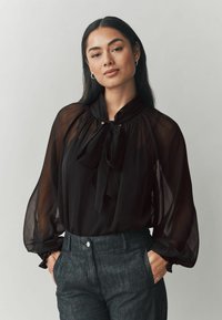 Čierna, priehľadná blúzka s vysokým golierom a detailom motýlika; má splývavé rukávy a voľný strih, kombinovaná s tmavými, presne strihanými nohavicami.