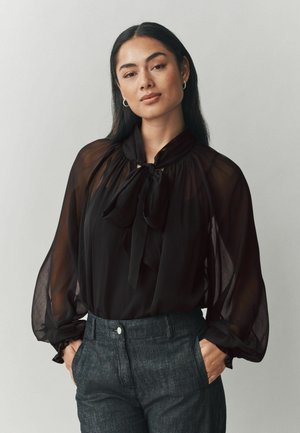 Blouse noire, transparente avec un col montant et des détails de nœud; présente des manches bouffantes et une coupe décontractée, associée à un pantalon sombre et ajusté.