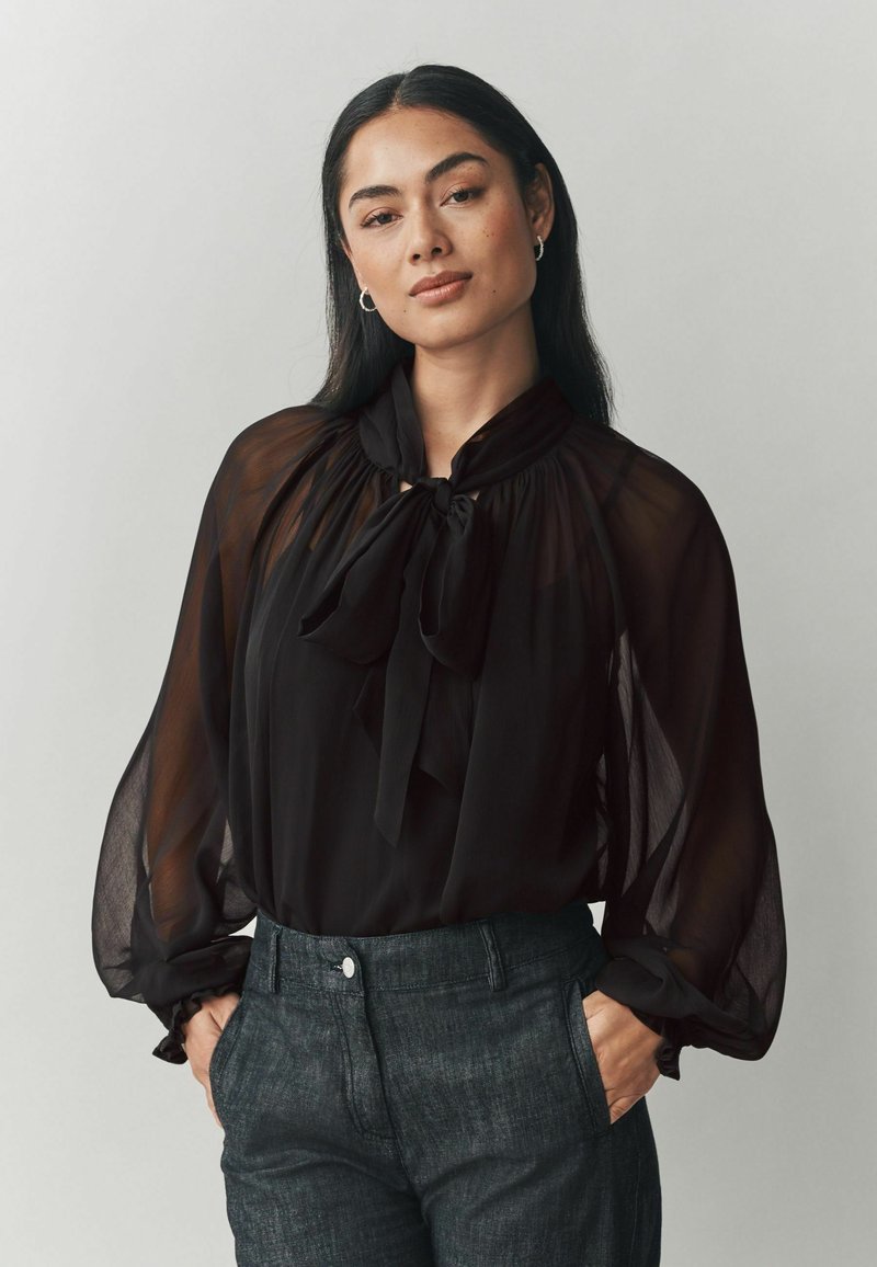 Čierna, priehľadná blúzka s vysokým golierom a detailom motýlika; má splývavé rukávy a voľný strih, kombinovaná s tmavými, presne strihanými nohavicami.