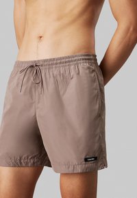 Costumi da bagno marroni realizzati in tessuto leggero e liscio. Presentano un elastico in vita con cordoncino e un piccolo logo nero applicato.