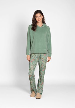 Woman standing wearing green hoodie, green floral patterned pants, and beige slippers against a plain light background.