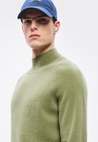 Zöld, puha anyagból készült magas nyakú pulóver, bordázott nyakkal és sima textúrával. Kék sapkával párosítva, amin logó látható.