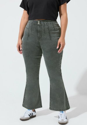 Person iført højtsiddende, falmede sorte flare-jeans, sort cropped t-shirt og hvide sneakers med sorte striber og blåt logo.