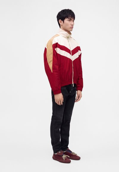 Homme debout portant une veste zippée rouge, blanche et beige, un jean noir et des baskets bordeaux sur un fond blanc uni.