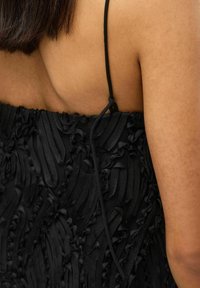 Juoda suknelė su tekstūruotu audiniu, puoštu sluoksniuotomis volanėlėmis. Plonos reguliuojamos petnešėlės ir užrišimo detalė nugaros pusėje. Matomos lygios odos.