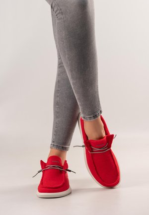 Person trägt graue, eng anliegende Jeans und leuchtend rote Freizeitschuhe mit hellgrauen Schnürsenkeln auf hellem, einfachem Hintergrund.