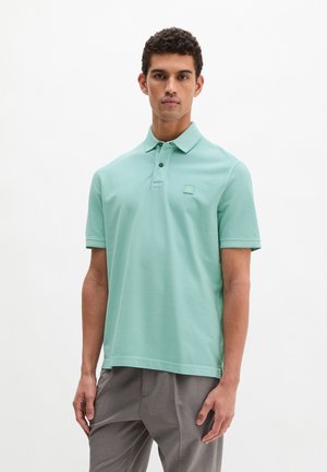 Uomo con polo a maniche corte azzurro chiaro e pantaloni grigi, in piedi contro uno sfondo bianco semplice, rivolto in avanti.