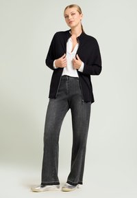 Zwarte zip-up jas met een relaxte pasvorm, gedragen over een wit shirt, gecombineerd met grijze wijde jeans en metallic sneakers. Zachte textuur, minimalistisch ontwerp.