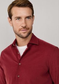 Rode overhemd met kraag, gemaakt van een gestructureerde stof. Het overhemd is over een wit T-shirt gedragen, waarbij subtiele bruine knopen zichtbaar zijn.