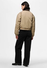 Cilvēks ar īsiem tumšiem matiem, kas ģērbts bežā bomber jakā, melnās platajās biksēs un melnās zābakos, stāv ar muguru pret vienkāršu fona.