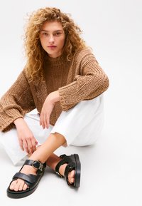 Hnedý pletený oversized sveter s širokými rukávmi, skombinovaný s bielymi nohavicami. Čierne sandále majú pracky a hrubú podrážku.