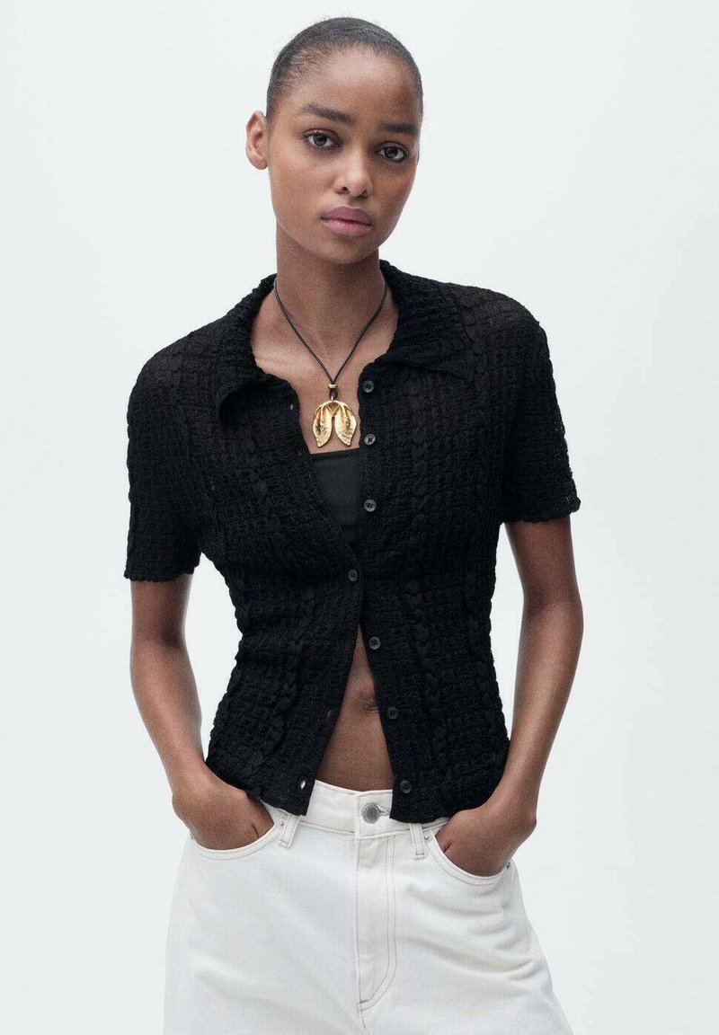 Chemise noire texturée à manches courtes avec un col et un boutonnage à l'avant, portée par-dessus un débardeur noir, associée à un jean blanc taille haute.