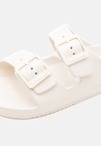 Hvide sandaler lavet af gummi med to justerbare remme med firkantede spænder. Tekstureret sål med brandnavn prentet på siden.