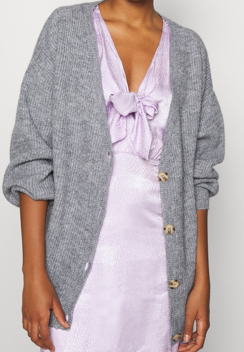 Gray knitted cardigan with oversized sleeves, featuring a row of beige buttons. Worn over a light purple dress with a bow detail.