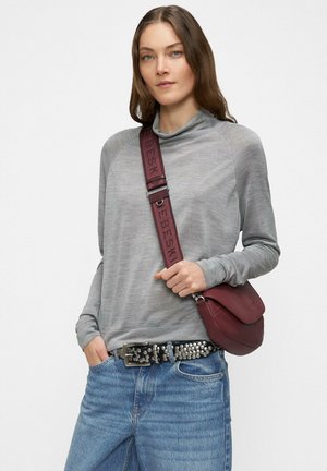 Woman wearing a grey long-sleeve top and blue jeans with a studded belt, carrying a burgundy crossbody bag with wide strap.