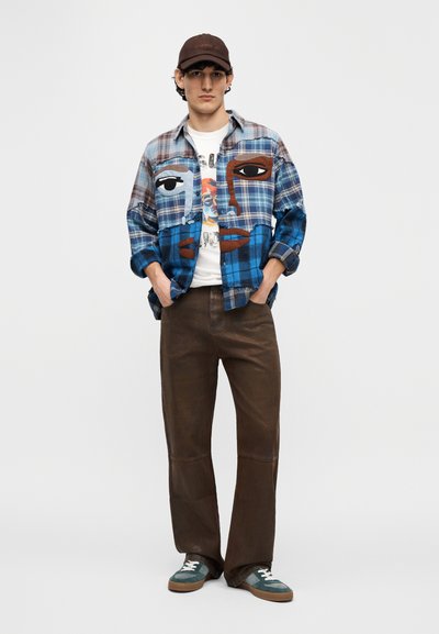 Πουκάμισο με καρό μπλε και καφέ τετράγωνα, μεγάλη εικόνα προσώπου, λευκό μπλουζάκι από κάτω, καφέ παντελόνι και σκούρα πράσινα αθλητικά παπούτσια. Φοράει καφέ καπέλο.