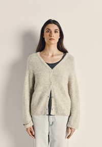 Heled halli, kohev kardigan V-kaelusega, nööbiga kinnitamine ja soonikkoes äär. Kandmise alla on valitud tumedat topi ja heledaid teksaseid.