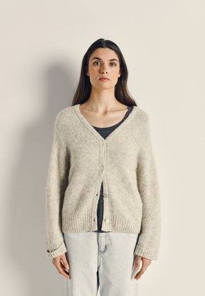 Cardigan gris clair et duveteux avec un col en V, fermeture à boutons et ourlet côtelé. Porté par-dessus un haut sombre et associé à un jean en denim clair.