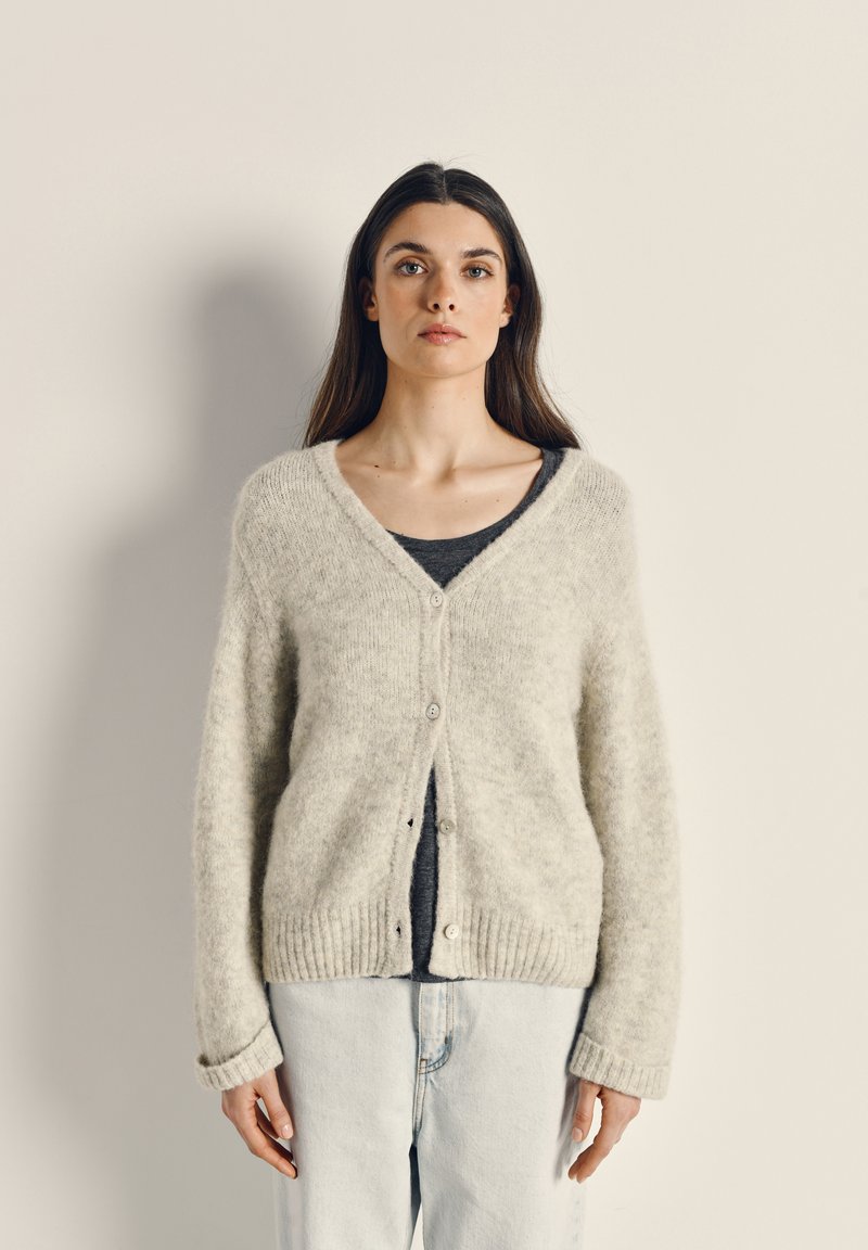 Heled halli, kohev kardigan V-kaelusega, nööbiga kinnitamine ja soonikkoes äär. Kandmise alla on valitud tumedat topi ja heledaid teksaseid.