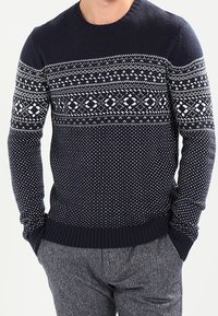 Mörkblå stickad tröja med vita geometriska mönster, med rund halsringning och ribbade muddar och fåll. Texturerad tygdesign.