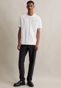 Witte katoenen t-shirt met korte mouwen en een subtiel logo op de borst, gecombineerd met donkergrijze rechtvallende jeans en zwarte nette schoenen.