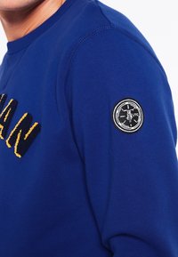 Sudadera azul con texto bordado en amarillo en la parte delantera. Cuenta con un parche circular negro en la manga izquierda con detalles en blanco.